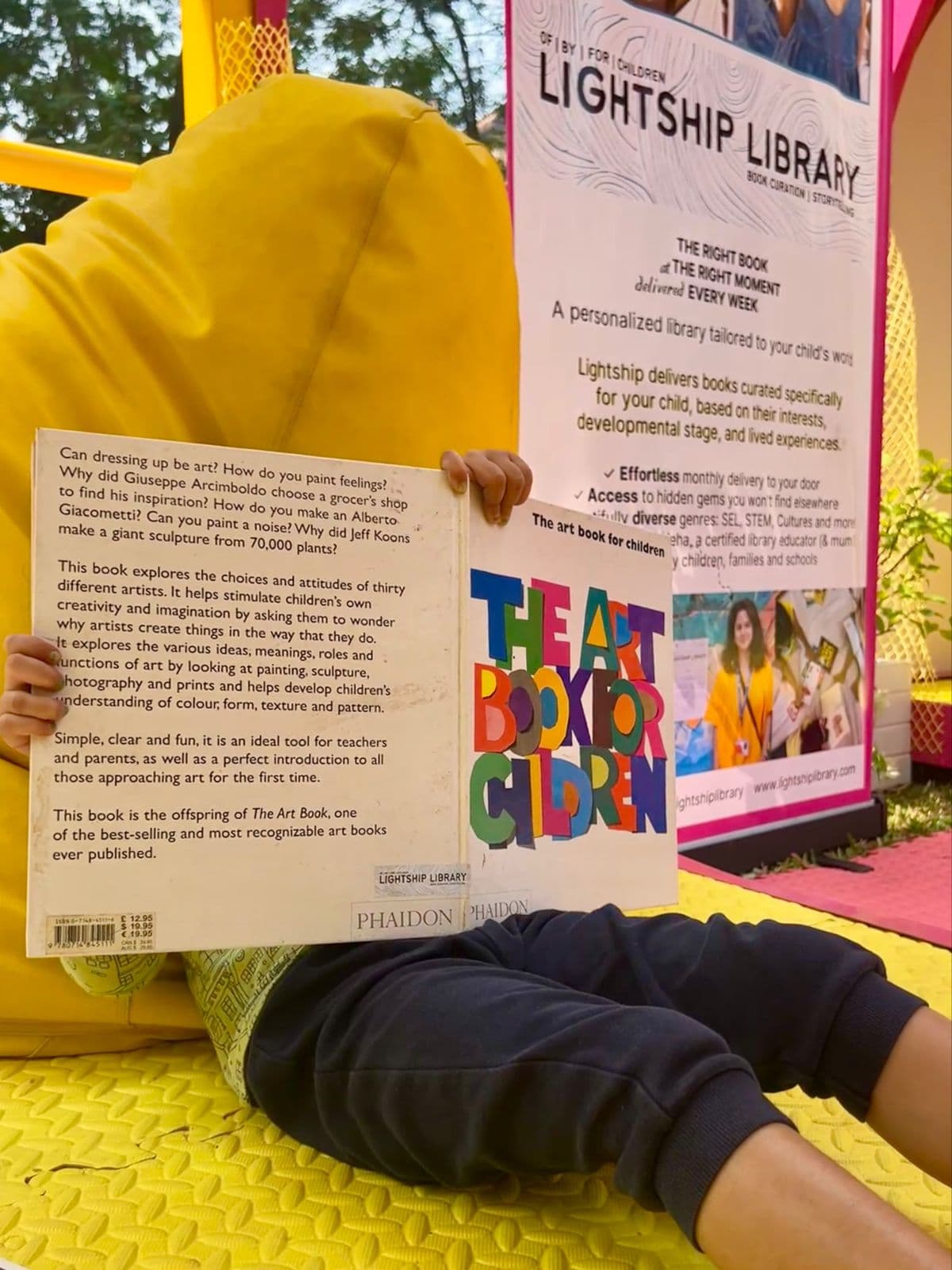 A child holding up a book beside the Lightship Library pop-up sign