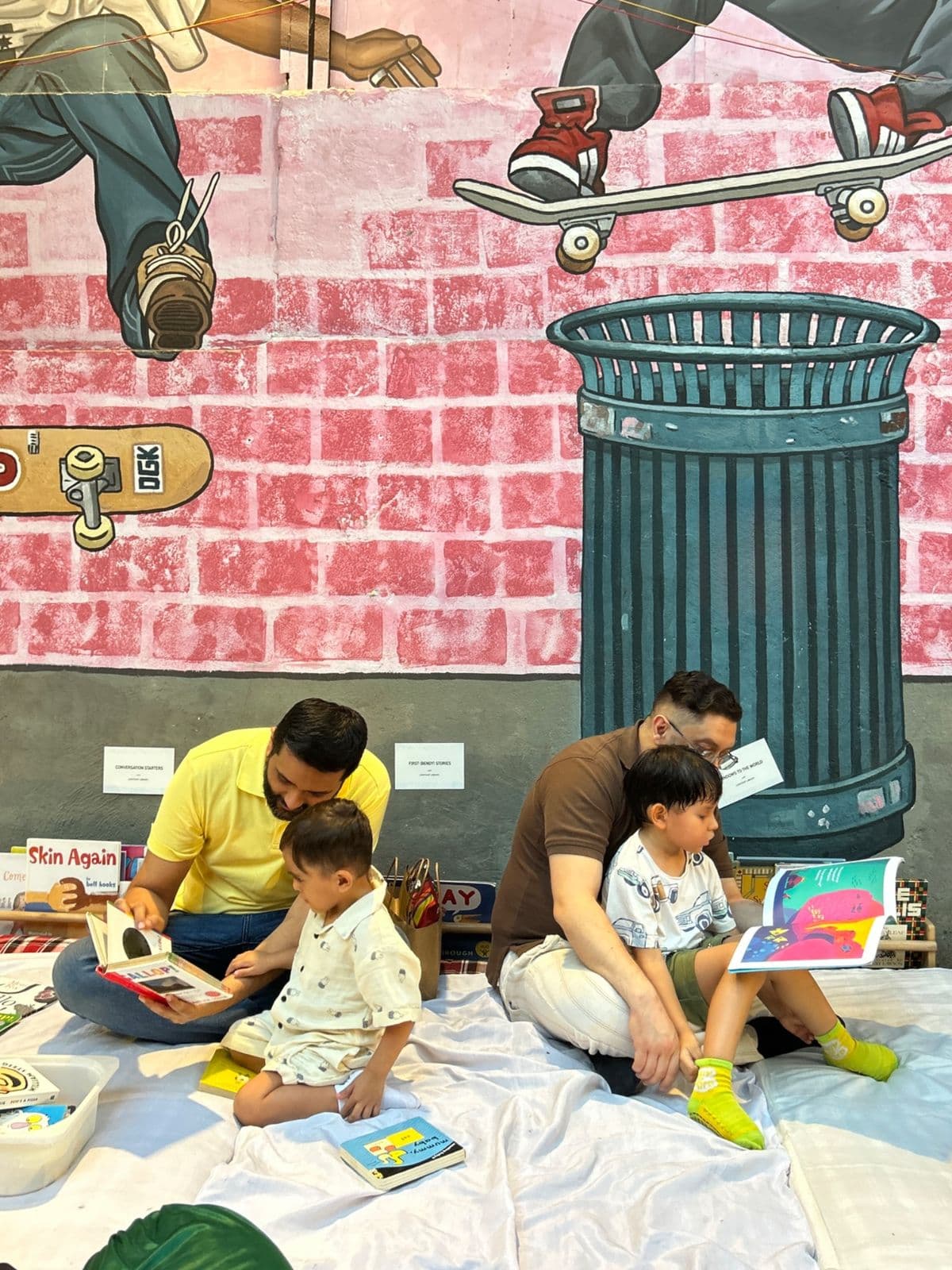Two fathers reading picture books with their toddlers on a blanket at a Lightship pop-up, in front of a painted brick-wall mural
